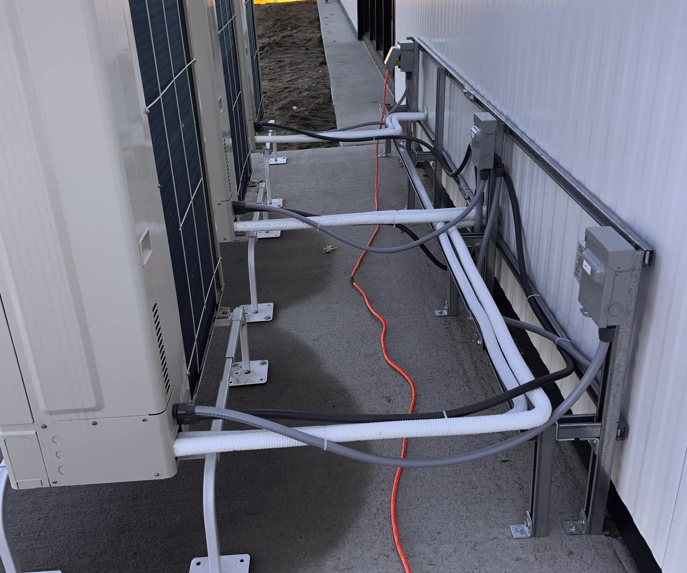 Außeneinheiten einer Wärmepumpe von hinten, davor gedämmte Leitungen und Anschlusskästen an der Wand