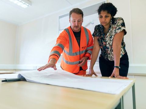 Ein Mann in Warnweste und eine Kollegin besprechen einen Plan auf dem Tisch